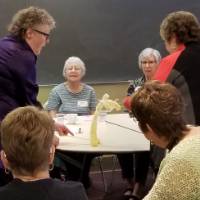A woman holding the sticks for a puppet's feet, a second woman holding the stick's for a puppet's arms, while 10 women sitting around a table look on